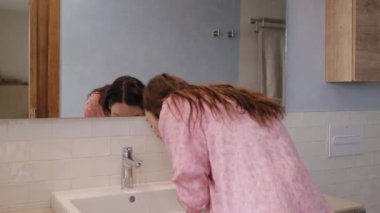 Pijamalı, gülümseyen genç bir kadının, banyoda makyaj aynasının önünde uzanırken yüzünü temiz suyla yıkaması. Sabah rutini. Güzel cilt bakımı konsepti. Video 4k yatay