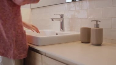Pijamalı tanınmayan bir kadın dişlerini temiz suyla fırçalamayı bitirir, lavaboya tükürür ve suyu kapatır. Sabah rutini. Yaklaş, yan görünüm, yatay 4k video