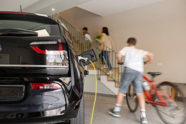 Belli bir garajda elektrikli bir aile arabasını şarj ederken, çocuk eve bisikletle ve aileyle birlikte merdivenlerden çıkarken. Hareket bulanıklığı, hareket, bulanık arkaplan, yatay