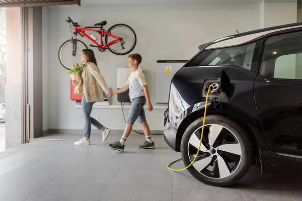 Happy family living in a home with an electric car in garage. Electric vehicle charging station in private home with blurred family leaving the house, with a bicycle hanging on the wall. Horizontal.