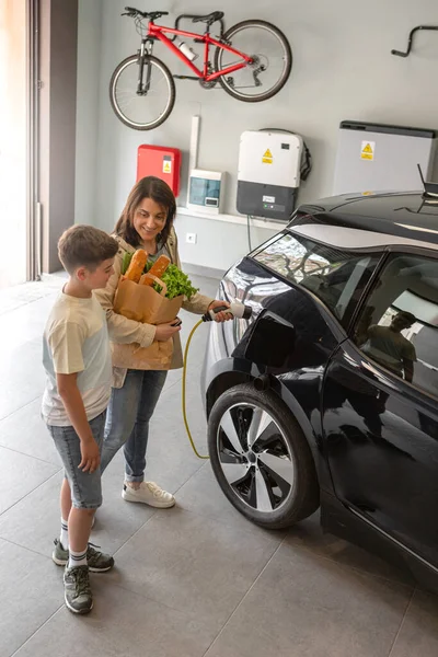 家庭用充電ステーションから電気自動車まで 幸せな息子と母親のプラグEv充電器 昼間に再生可能エネルギーを動力とする未来の環境に優しい車 高角度 — ストック写真