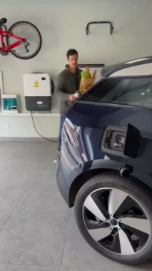 Adam marketten alışveriş yaptıktan sonra evdeki garajda şarj aletini elektrikli mavi araba prizine takıyor. Yenilenebilir evler, Enerji Verimli Ev Hayatı. Dikey Video 4k 'yi kapat.