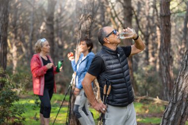 Bir grup kıdemli arkadaş orman yürüyüşü sırasında su içmek için durdular, ellerinde su şişeleri ve yürüyüş direkleriyle, açık havanın tadını çıkardılar ve doğada aktif kaldılar. Su içen adam. Yatay
