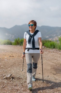 Gülümseyen yaşlı bir kadın güneş gözlüğü ve spor kıyafetleriyle bir tarafa bakıyor, kirli bir yolda sırıklarla yürüyor. Yeşil çalılar ve dağlarla çevrili, ve güneş ışığı bulutlu gökyüzü. Dikey