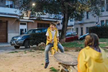 İki çocuk, akşamları mahalledeki bir parkta tahterevallide eğleniyor. Doğal neşe ve etkileşim, hafif ışıklandırmayla orta ölçekte çerçevelenmiş. yatay kopyalama alanı