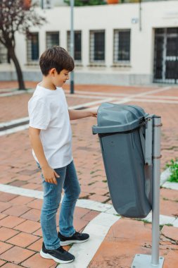 Beyaz tişörtlü ve kot pantolonlu genç bir çocuk halka açık bir çöp kutusuna çöp atıyor. Temiz ve sorumlu bir atmosferi olan şehir merkezi. Çevresel farkındalık kavramı. Dikey kopyalama alanı