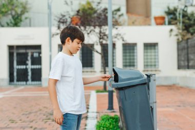 Beyaz tişörtlü ve kot pantolonlu genç bir çocuk halka açık bir çöp kutusuna çöp atıyor. Temiz ve sorumlu bir atmosferi olan şehir merkezi. Çevresel farkındalık kavramı. Yatay kopyalama uzayı