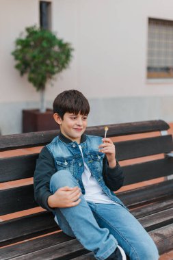 Günlük kıyafetli bir çocuk açık hava bankında oturuyor, bir lolipopla muzip bir ifadeyle gülümsüyor. Şehir merkezi, doğal ışık, sakin bir yerleşim alanı. Dikey, kopyala