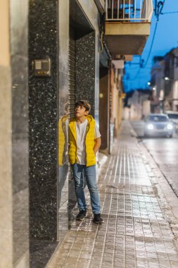 Sarı yelekli bir çocuk kapının önünde duruyor, zili çalıyor ve bekliyor. Sıcak aydınlatma, park edilmiş arabalar ve sakin bir şehir atmosferi olan huzurlu bir şehir caddesi. Dikey kopyalama alanı