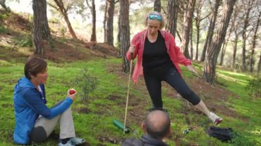 Bir grup kıdemli yürüyüşçü yürüyüş gününden sonra bir orman açıklığında mola verir. Bir kadın esnerken, diğerleri meyve ve suyla kendilerini tazeler. Yuvarlak izleme çekimi, doğal ışıklandırma..