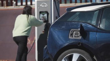 Yeşil kazaklı bir kadın elektrik teçhizatı makinesinden mavi elektrikli bir arabanın arkasına güç kablosunu bağlıyor. Sürdürülebilir enerji kullanımı kavramı. Yatay, 4k 