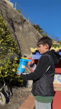 Kendinden emin çocuk bahçesinde sprey şişesiyle böcek ilacı gübresi uyguluyor. Bitkilerin büyümesine yardım etmeye odaklanmış, mavi gökyüzü ve arka planda yeşil yapraklarla, sorumluluklarını gösteriyor