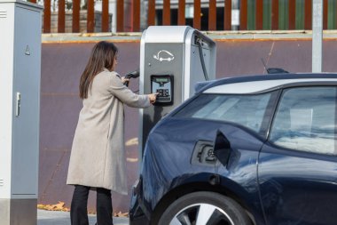 Bir kadın arabasının fişini takarken bir elektrikli araç şarj istasyonunu çalıştırıyor, temiz enerjiyi teşvik ediyor. Modern şehir geçmişi. Kopyalama alanı ile yatay ortam görüntüsü.