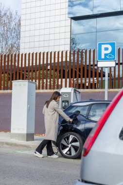 Kadın elektrikli aracını EV istasyonunda şarj ediyor. Arka plandaki modern ofis binaları kentsel sürdürülebilirliği ve çevre dostu ulaşımı vurguluyor. Geniş açı, dikey kopyalama alanı.