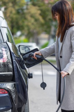 Tanınmayan bir kadın elektrikli arabasını bir şehir caddesinde halka açık bir şarj istasyonuna takıyor. Çevre dostu ulaşım ve sürdürülebilir enerji kavramı. Yakın çekim, dikey perspektif.