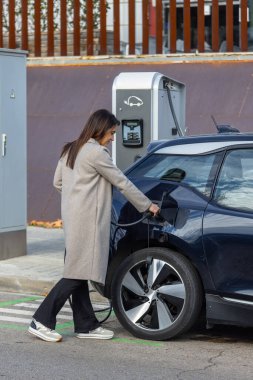 Bir kadın elektrikli arabasını gündüz vakti bir şehir caddesinde bir şarj istasyonunda şarj ediyor. Sürdürülebilir hareketlilik ve çevre dostu ulaşım konsepti. Yan açı, Dikey