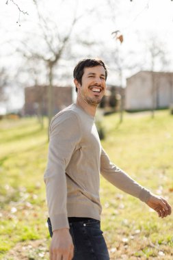 Neşeli bir yüzü olan, güneşli bir parkta yürüyen sıradan giyinmiş bir adam. Bej renkli bir kazak ve bulanık bir arka plan, ağaçlar, çimenler ve binalardan oluşan kot pantolon giyiyor. Kopyalama alanı ile dikey