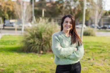 Yeşil kazak giyen kendine güvenen bir kadın kolları çaprazlanmış, kameraya gülümseyen bir şehir parkında duruyor. Doğal gün ışığı, açık hava portresi, şehir arka planı. Yatay bileşim, kopyalama alanı.
