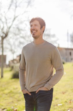 Gülümseyen orta yaşlı bir adam dışarıda duruyor, doğadaki güneş ışığının tadını çıkarıyor. Rahat duruş ve sıcak ifade pozitifliği ve refahı iletir. Ağaçlar ve binalarla dikey, yumuşak odaklı arkaplan