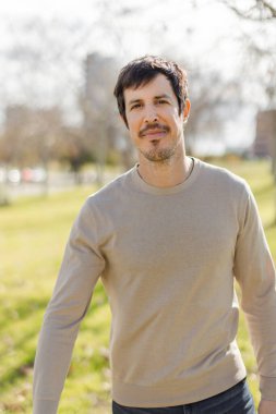 Hafif sakallı, orta yaşlı, kendine güvenen yakışıklı bir adam güneşli bir parkta hafifçe gülümsüyor. Kameraya bakıyorum. Bulanık şehir arka planı ve yeşil çimenler. Dikey, yumuşak odak, doğal ışık