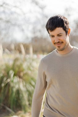 Orta yaşlı beyaz bir adam, güneşli bir parkın içinden nazik bir gülümsemeyle aşağıya doğru yürür. Düşünce ve barış kavramı. Dikey, bulanık yeşillik, yumuşak doğal ışık, ve boşluğu kopyala.