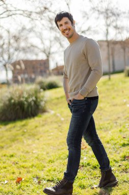 Orta yaşlı beyaz bir adam yeşil bir parkta yürüyor, temiz havanın tadını çıkarırken gülümsüyor. Kendinden emin adımlar ve rahat tavırlar. Tasasız ve olumlu bir bakış açısı. Dikey, bulanık şehir arkaplanı 