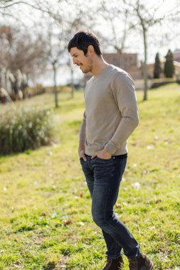 Orta yaşlı beyaz bir adam güneşli bir parkın içinden geçer, barışçıl ve doğal bir ifadeyle yan tarafa bakar. Kaygısız bir an. Tüm gövde çekimi, dikey, bulanık arka plan, ağaçlar ve çimenler.