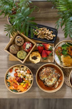 Taze yemek, salata ve ahşap bir masada, çevre dostu kâselerdeki tatlıları tepeden tırnağa görmek. İçinde kurabiye, peynirli kek, salata ve taze meyve var. Yeşil bitkiler resmi çerçeveler, doğal dokunuş.