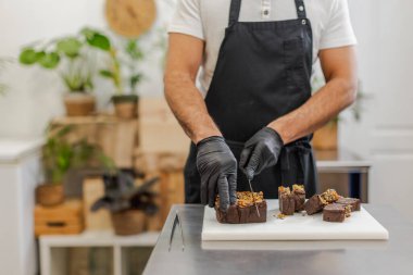 Tanınmayan pasta şefi, kısmen görünür, paslanmaz çelik masadaki beyaz bir kesim tahtasında çikolata tatlısı dilimliyor. Yatay, bulanık arkaplan, kopyalama alanı.
