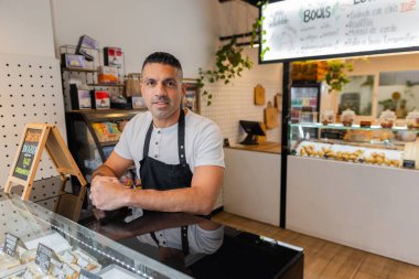 Siyah önlüklü, dost canlısı bir fırın sahibi cam tezgahın üzerine eğiliyor ve çeşitli hamur işleri sergiliyor. Sıcak ortam, modern dükkan içi, küçük işletmeler. Yatay ve kopyalama uzayı