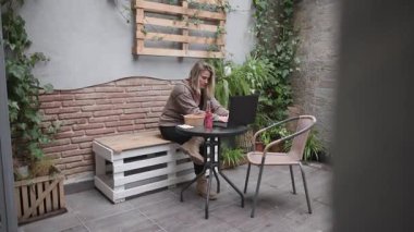 Bir kadın, rahat bir kafe terasında dizüstü bilgisayarı üzerinde çalışırken çevre dostu bir kaseden yemek yiyor. 4k video.