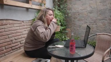 Kırsal bir kafede oturan bir kadın laptopuyla çalışırken çevre dostu bir kaseden yemek yiyor. Masada yeşilliklerle çevrili bir içecek ve çatal bıçak takımı var. Orta ekran, kenar görüş.