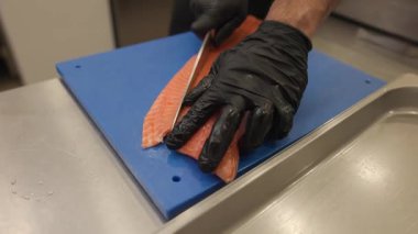 Bir şefin büyük taze bir somon filetosunu mavi bir kesme tahtasıyla ayrı ayrı keserken yakın çekimi. Porsiyonlar daha sonra pişirmek için bir tepsiye konur. Ufak tefek iş hazırlıkları. 4k video