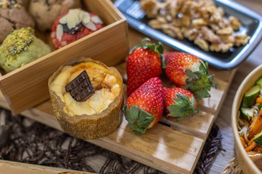 Arkaplanda ceviz tepsisi ve salata kasesi olan ahşap bir tahtanın üzerinde zanaatkâr keklerin, kurabiyelerin ve taze çileklerin yakın çekimi. Doğal ışık altında renkli yemek tasarımı. Yatay ve kopyalama uzayı.