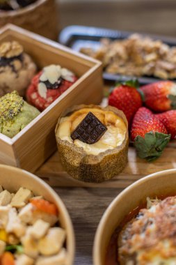 Koyu çikolatayla kaplı mini bir peynirli kekin dikey yakın çekimi. Tatlı, taze çileklerin ve çeşitli kurabiyelerin arasında konforlu, kırsal bir yiyecek tasarımı düzeninde hazırlanır. Gurme servis yemeği..