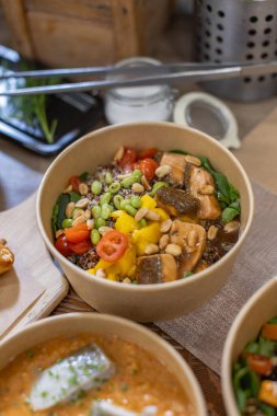 Ispanak üzerine jöleli somon, mango parçaları, vişneli domates, edamame, fıstık ve kinoa içeren canlı bir poke kasesi. Tahta masaya dikey yakın çekim. Paket servisi, çevre dostu ambalaj