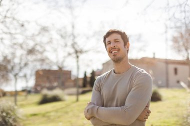 Kendine güvenen bir adam kollarını kavuşturup güneşli bir parkta kameraya bakarken nazikçe gülümser. Sakinliği ve güveni, refahı, kendini geliştirmeyi, koçluğu ya da yaşam tarzını gösterir. Doğal ışık