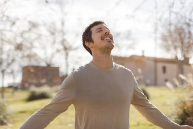 Gözleri kapalı bir adam güneşe bakarken hafifçe gülümser. Huzuru, minnettarlığı, zihinsel berraklığı ifade eder. Sağlık, denge ya da farkındalık temaları için mükemmel. Yatay, bulanık park arkaplanı.