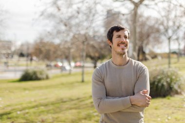 Aydınlık bir günde dışarıda duran bir adam, kollarını kavuşturmuş, yumuşak bir gülümseme, rahat bir duruş. Özgüveni, sıcaklığı, kişisel gelişimi çağrıştırıyor. Yatay, doğal ışık, şehir parkı bulanık arka plan. Kopyalama alanı