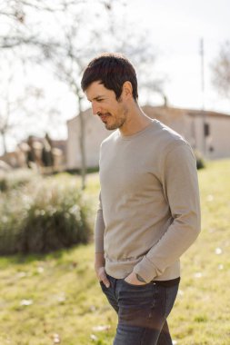 Elleri ceplerinde sıradan bir adam parkta yürüyor, yumuşak bir ifadeyle aşağıya bakıyor. İçgözlem, sakinlik ve farkındalık gösteriyor. Dikey, doğal ışık, yumuşak arka plan bulanıklığı.