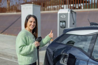 Yeşil kazaklı mutlu bir kadın elektrikli araba şarj aleti tutuyor ve onaylıyor. Arka planda kentsel EV şarj istasyonu.