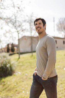 Elleri ceplerinde, yumuşak bir gülümsemeyle parkta yürüyen rahat bir adam. Rahatlık, güven ve doğal cazibeyi ifade eder. Yaşam tarzı, sağlık ya da modern erkeklik teması için ideal. Dikey, kopyala.