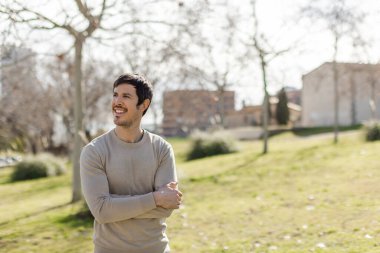 Güneşli parkta duran, gülümseyen ve mesafeye bakan bir adam. Rahatlamış ifadesi iyimserliği, refahı ve özgürlüğü gösteriyor. Sağ tarafında temiz kopyalama alanı olan yatay resim.