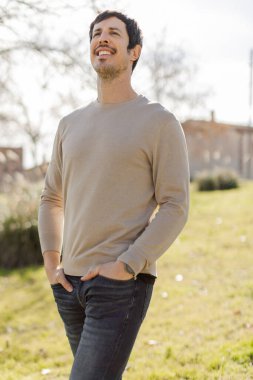 Elleri ceplerinde güneşli bir parkta yukarı bakıp gülümseyen bir adamın portresi. Umudu, minnettarlığı ve varlığı iletir, sağlık, yaşam tarzı veya kişisel gelişim projeleri için idealdir. Dikey.