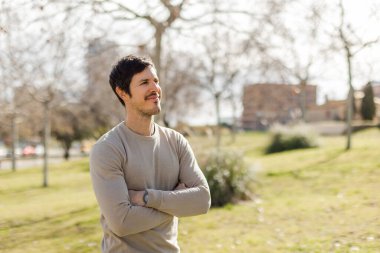 Kollarını kavuşturmuş düşünceli genç bir adam aydınlık bir günde uzaklara bakıyor. Huzurlu bir ifade, bej süveter, yumuşak ışık. Yansıma, gelecek görüşü, sakinlik. Kopyalama alanı ile yatay.