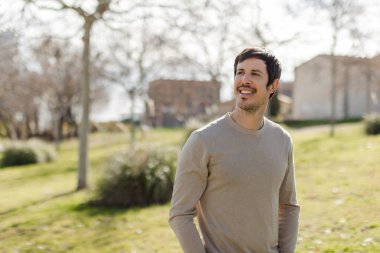 Parkta yürüyen bir adam, mesafeye bakarken usulca gülümsüyor. Sıradan tarz, rahat duruş. İyimserliği, kişisel gelişimi ve yaşam tarzı dengesini gösteriyor. Kopyalama alanı ile yatay.