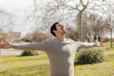 Adam kolları açık, gözleri kapalı, yüzünde güneşi hissederek hareketsiz duruyor. Sakinlik, minnettarlık ve akıl sağlığını gösteriyor. Yaşam tarzı, denge ya da kişisel bakım temalarına uyuyor. Yatay, yumuşak doğal arkaplan.