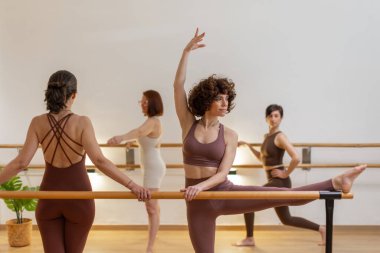 Dört kadın, ahşap zeminli ve yumuşak ışıklandırmalı sıcak tonlu bir stüdyoda bale barı yapıyorlar. Barda bacak esnetme hareketi. Uyumlu atmosfer gücü, zarafeti ve kontrolü harmanlıyor. Yatay.