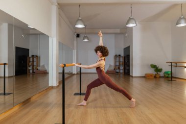 Odaklanmış bordo giysili bir kadın geniş stüdyodaki barda aynalar, ahşap zemin, kolye ışıkları ve yeşil bitkilerle güç ve esneklik pozu veriyor. Yatay, kopyalama uzayı