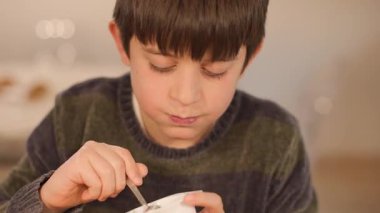 Annesi arka planda hareket ederken rahat ve neşeli bir yüzle doğal yoğurt yiyen 10 yaşındaki bir çocuğun yakın çekimi. Sakin, her gün. Yatay, 4K